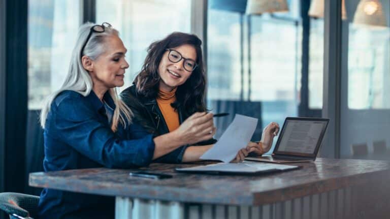 Deux femmes discutant au bureau, assises devant des feuilles de papier. L'une d'elle a les cheveux blancs et des lunettes dans les cheveux, l'autre les cheveux bruns et porte des lunettes.