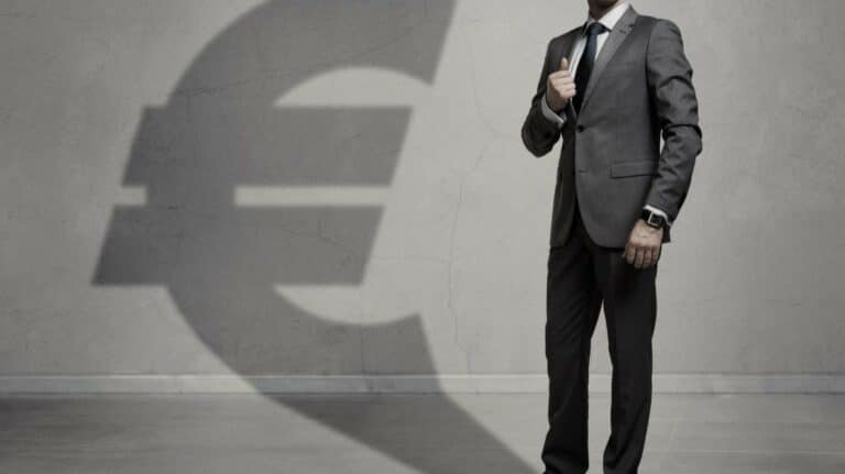 Un dirigeant d'entreprise en costume projetant une hombre en forme d'euro sur un mur gris.