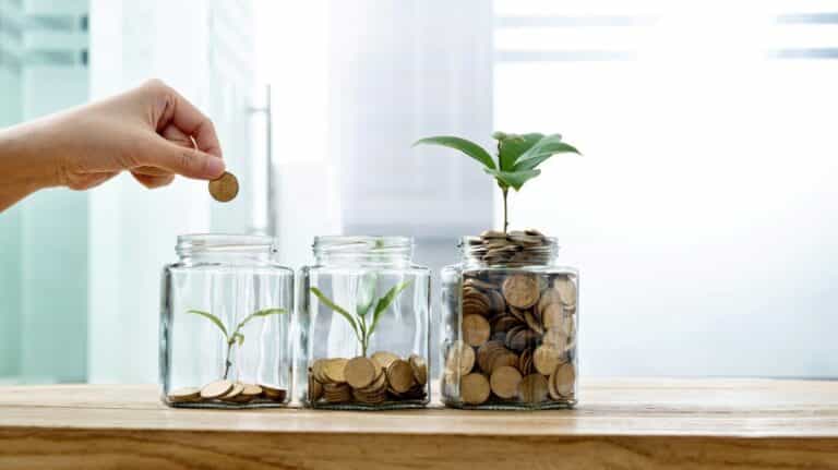 3 pots en verre remplis de pièces avec une plante qui pousse dans chacune d'elles. Une main dépose une pièce dans le pot à gauche.