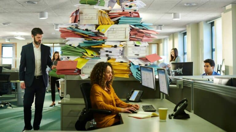 Une femme travaillant à son bureau pendant qu'une pile de dossiers s'amoncelle derrière elle