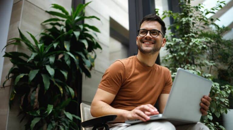 Un jeune homme travaillant sur un ordinateur, il est assis entre 2 plantes