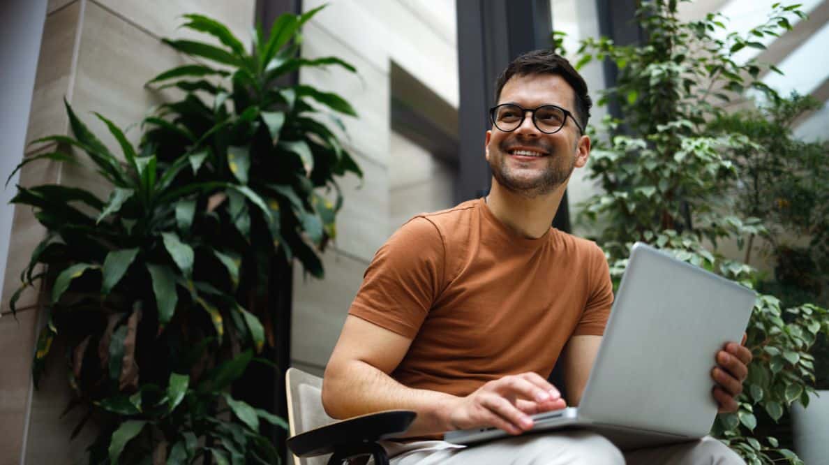 Un jeune homme travaillant sur un ordinateur, il est assis entre 2 plantes