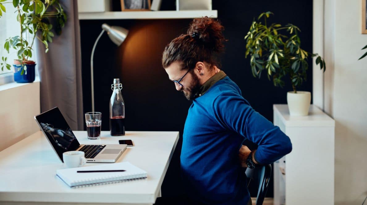 Un homme se cambre car il a mal au dos. Il est assis à son bureau derrière son ordinateur portable.